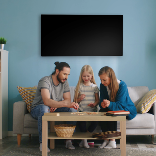 A family of three sitting on a sofa playing dominos with a black coloured Yandiya far-infrared heating panel behind them installed high on the wall